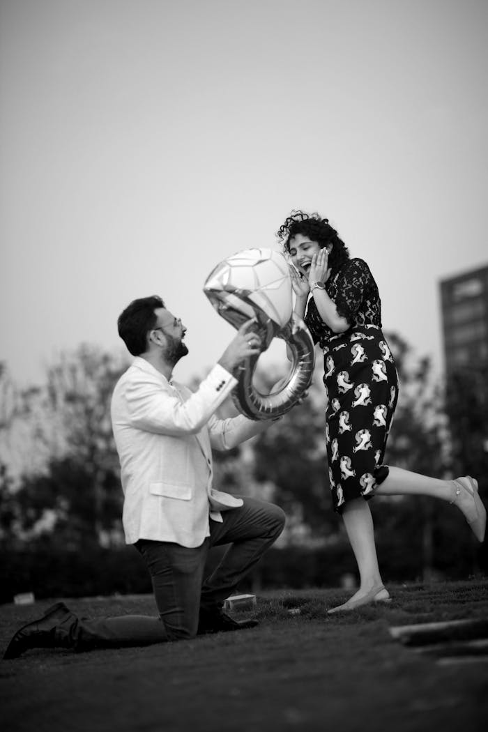 A man proposes to a woman with balloons in a park setting, captured in black and white.
